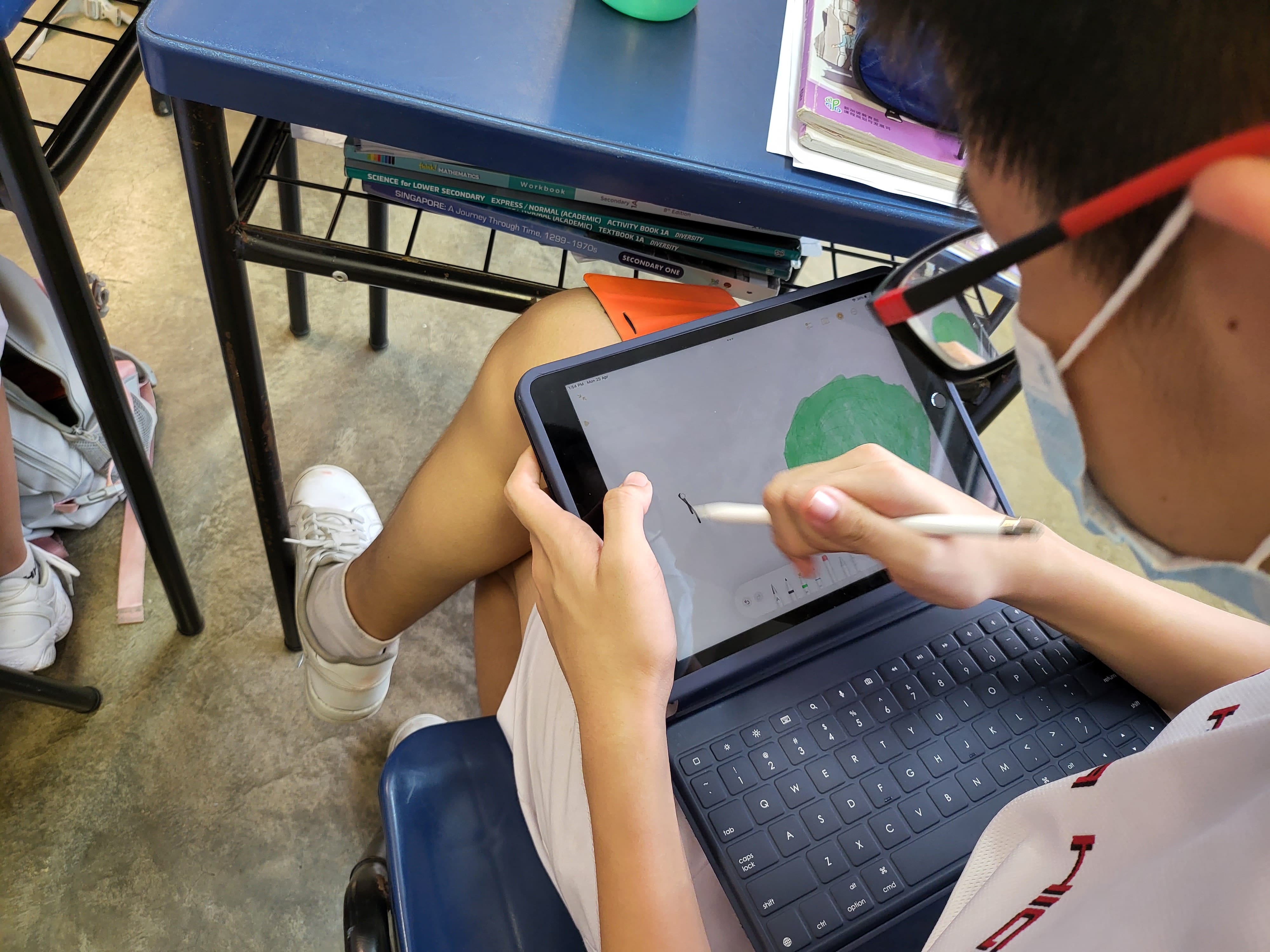 A masked student draws on an iPad with a stylus, seated near a desk stacked with school textbooks.